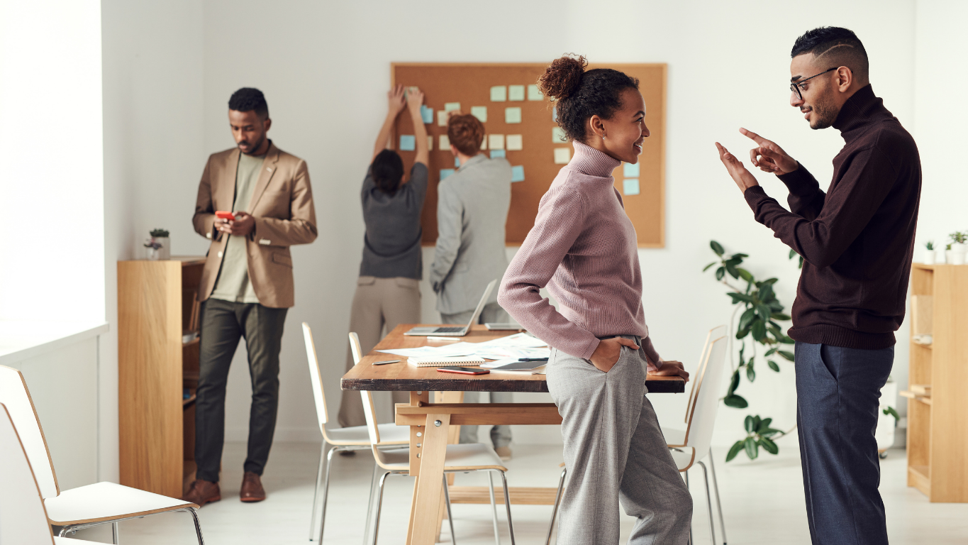 Two people talking in office with people working in background